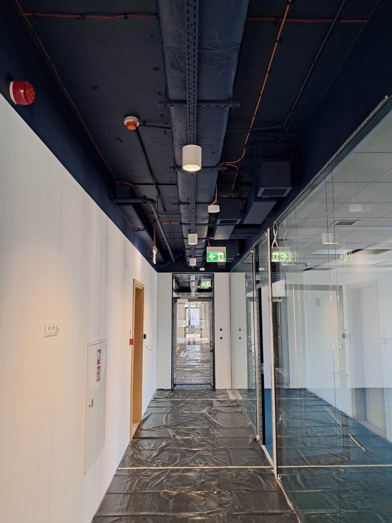 Korytarz biurowy w trakcie prac wykończeniowych: zabezpieczona podłoga folią, widoczne instalacje pod sufitem, oświetlenie punktowe, czujniki dymu i tabliczki z kierunkiem ewakuacji.