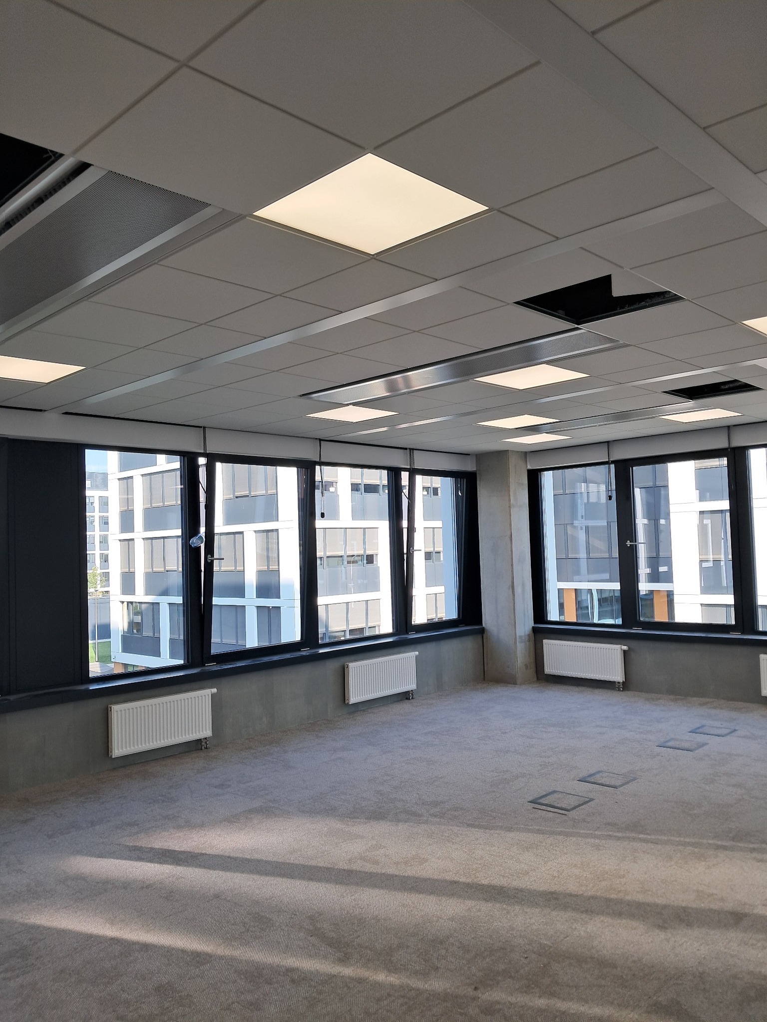 Wnętrze biurowe z oknami, widoczne grzejniki, podwieszany sufit z panelami oświetleniowymi i otworami wentylacyjnymi, szara wykładzina dywanowa.