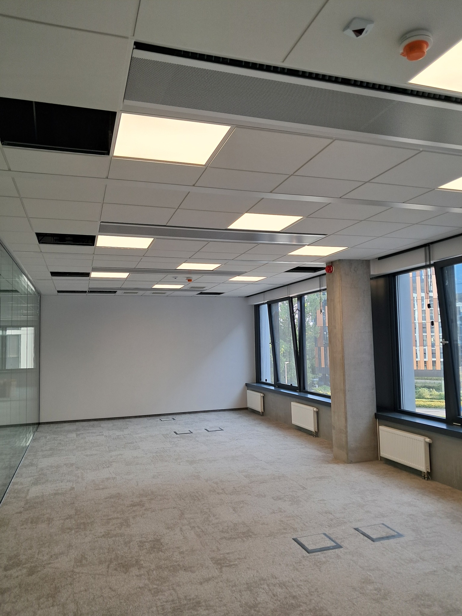 Wnętrze biurowe w stanie deweloperskim z widocznymi panelami sufitowymi z oświetleniem LED, instalacją przeciwpożarową, grzejnikami pod oknami i wykładziną dywanową, przygotowane do dalszego...