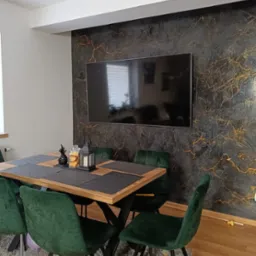 Eleganckie wnętrze jadalni z dekoracyjną ścianą w odcieniach szarości i złota, na której wisi telewizor. Stół z ciemnymi podkładkami i dekoracjami, otoczony zielonymi, welurowymi krzesłami. Podłoga...