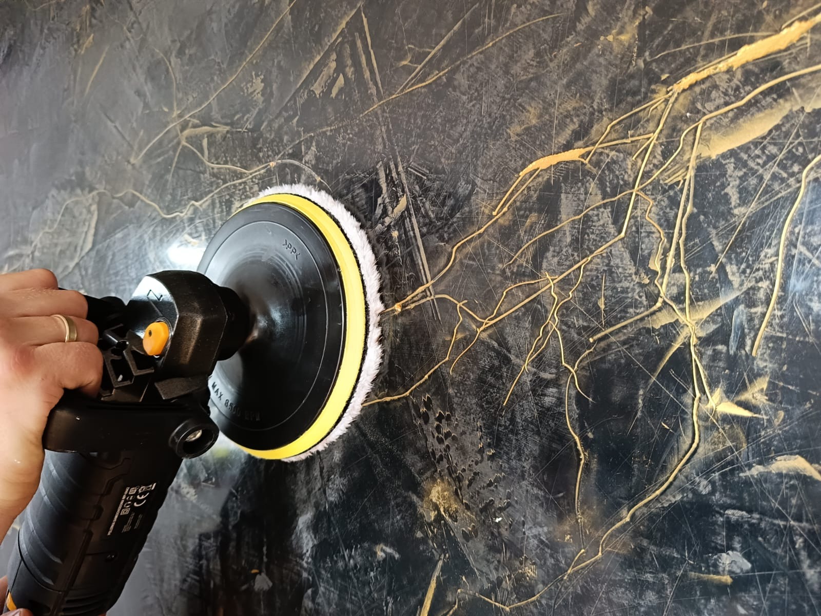 Polerowanie dekoracyjnej ściany w odcieniach czerni i złota za pomocą polerki elektrycznej z białym futrem, widoczny fragment dłoni z obrączką.