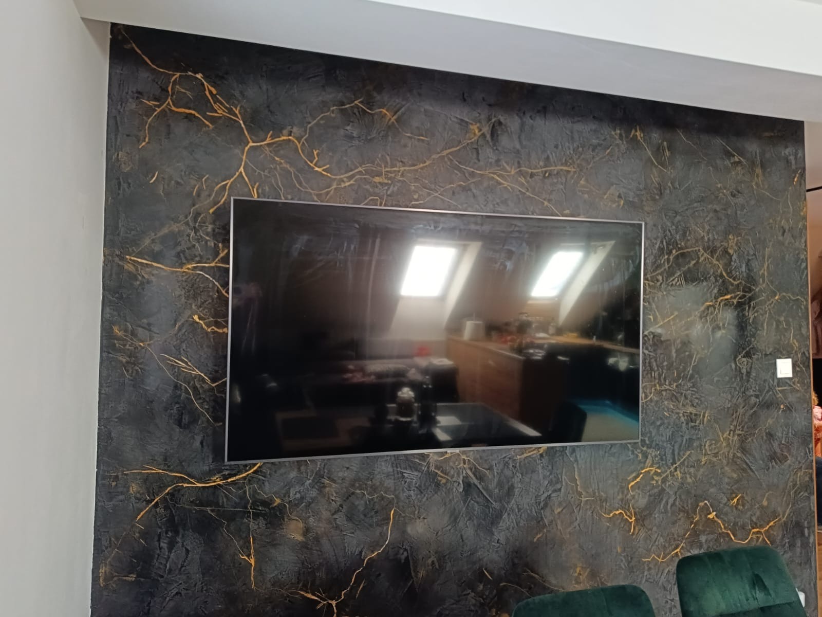 Ściana w salonie z dekoracyjną, ciemną strukturą imitującą żyły złota, na której zawieszony jest telewizor odbijający wnętrze pokoju na poddaszu.