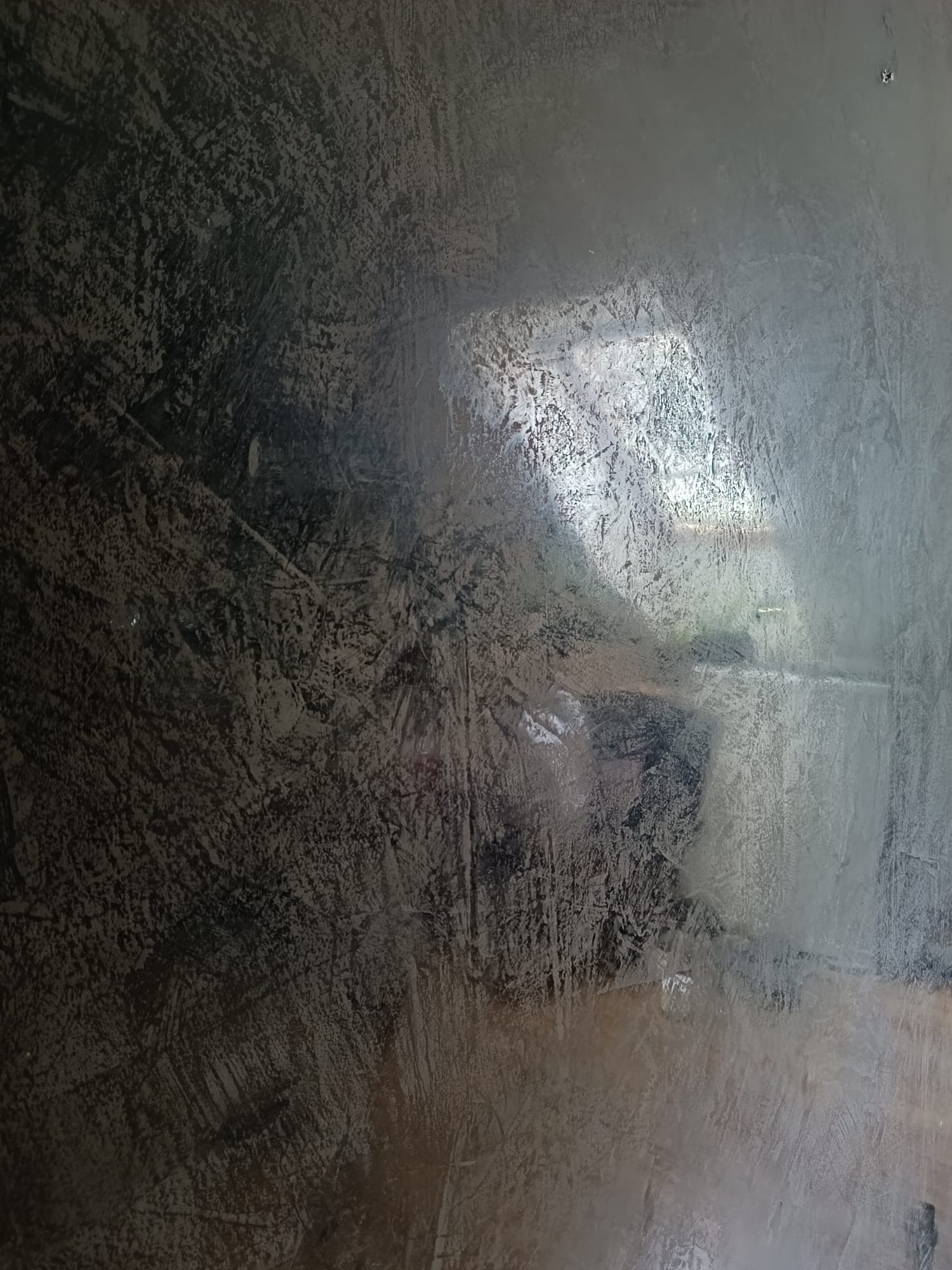 Nierównomiernie nałożona, ciemna, strukturalna powierzchnia z widocznymi smugami i odbiciem światła, przypominająca surowy beton lub niedokończone szpachlowanie.