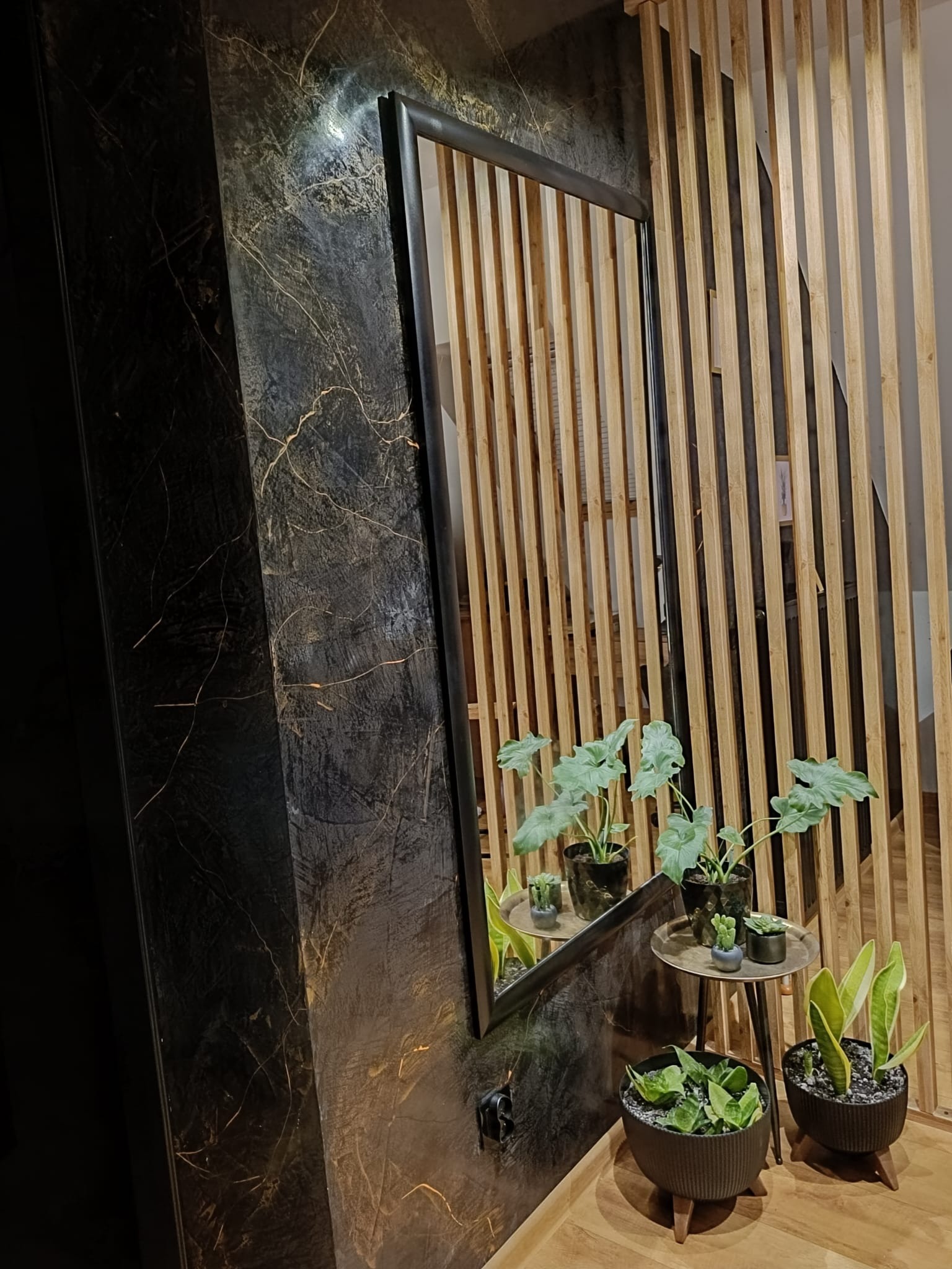 Ściana wykończona czarnym tynkiem dekoracyjnym z subtelnymi złotymi żyłkami, obok drewniana ażurowa ścianka działowa i lustro w czarnej ramie odbijające rośliny doniczkowe.