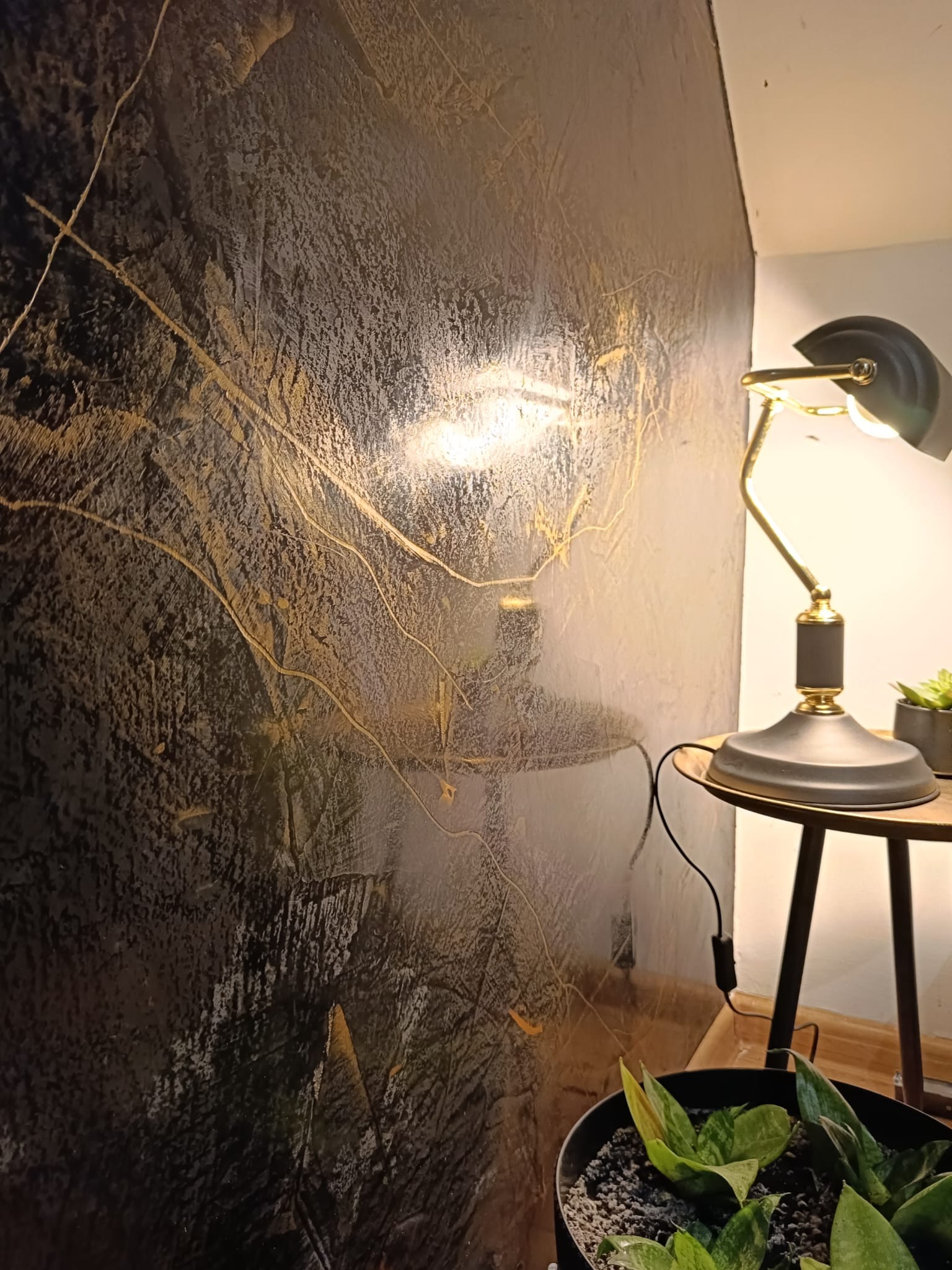 Dekoracyjna ściana z efektem przetarć w odcieniach szarości i złota, oświetlona lampką nocną na stoliku, obok doniczka z rośliną.