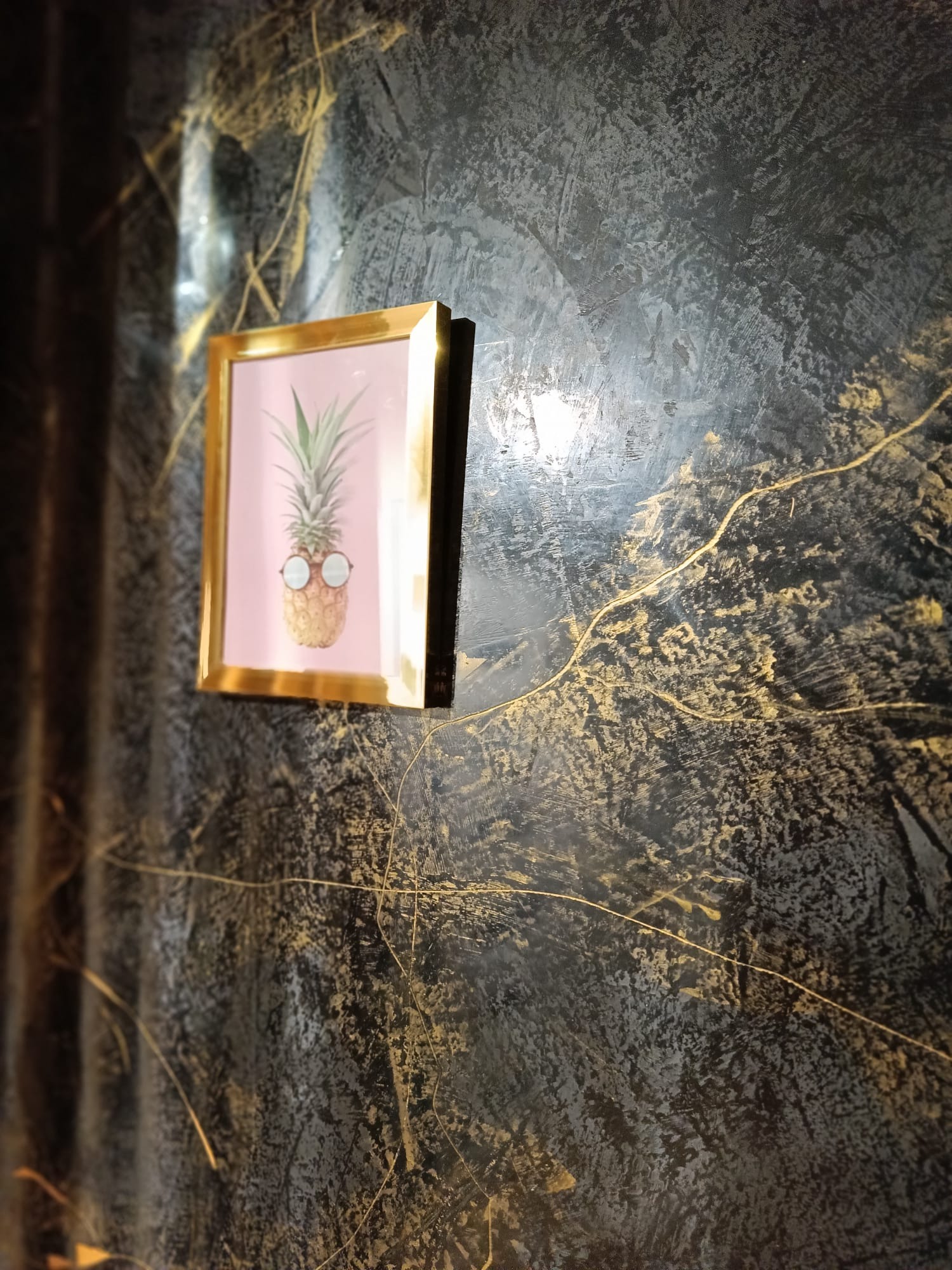 Dekoracyjna ściana w odcieniach szarości i złota, z efektem strukturalnym i zawieszonym obrazkiem ananasa w okularach w złotej ramce.