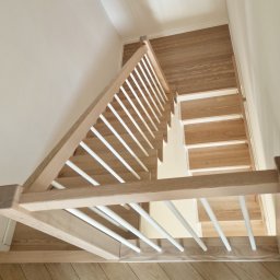 JB stolarnia-producent schodów na wymiar - Nowoczesne schody z jasnego drewna o widocznym usłojeniu, z minimalistyczną balustradą z białymi, cienkimi tralkami. Ujęcie z góry, ukazujące konstrukcję i podest.