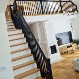 JB stolarnia-producent schodów na wymiar - Schody dębowe z białymi podstopnicami i czarną balustradą z toczonymi tralkami, prowadzące na antresolę. W tle salon z kaflowym kominkiem i parkietem w jodełkę.