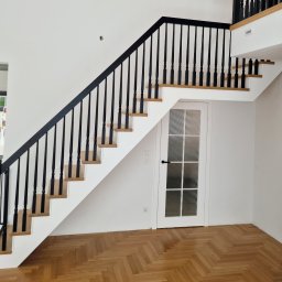 JB stolarnia-producent schodów na wymiar - Nowoczesne schody z czarną balustradą i jasnymi, drewnianymi stopniami, osadzone na białej konstrukcji. Pod nimi białe drzwi z matowymi szybami. Na podłodze parkiet w jodełkę.