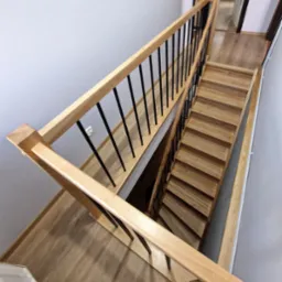 Jasne, dębowe schody z czarnymi, metalowymi balustradami, widok z góry, z drewnianą podłogą i białymi ścianami.