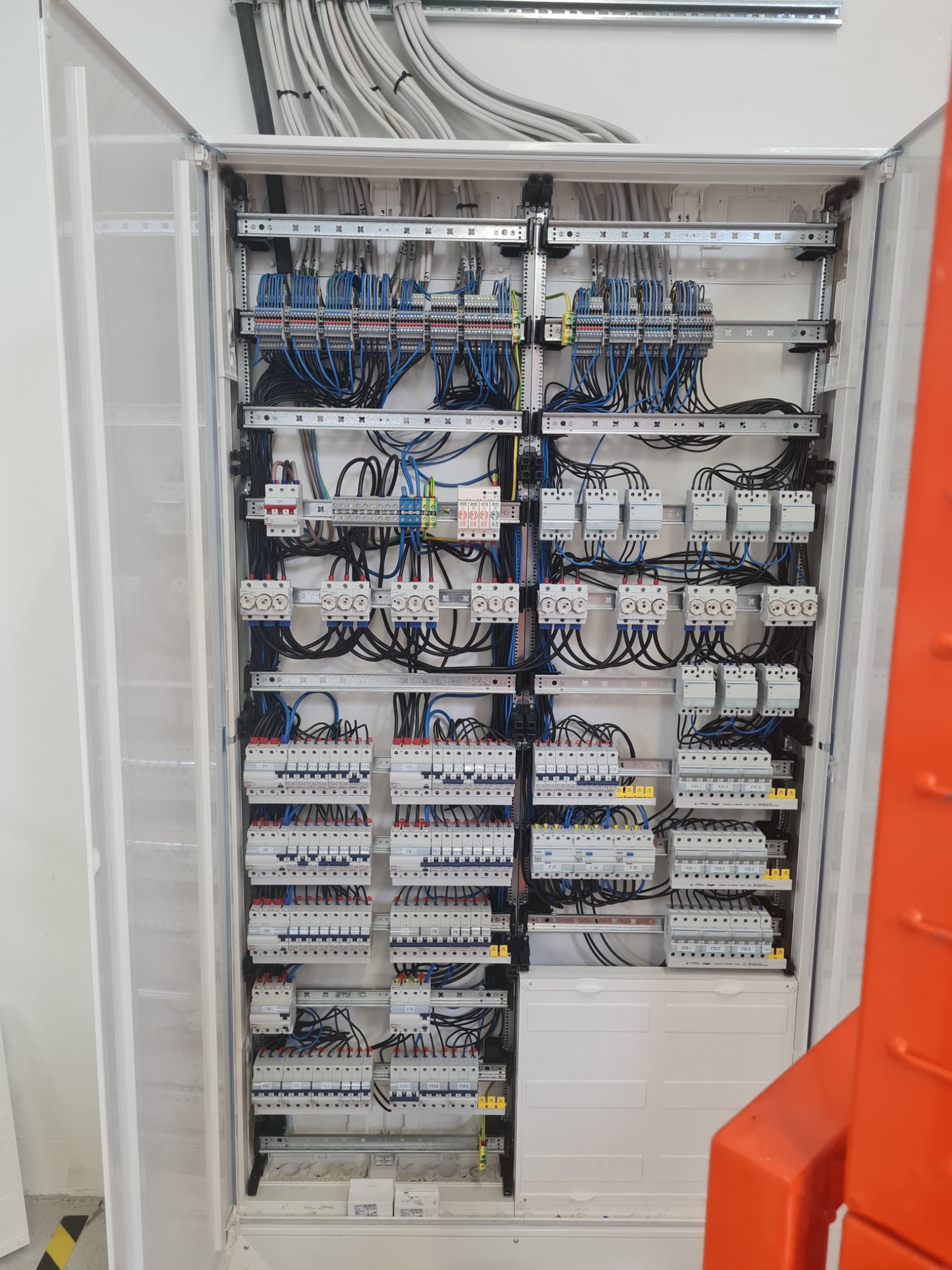 Otwarta szafa rozdzielcza z licznymi zabezpieczeniami, bezpiecznikami i przewodami, starannie ułożonymi i podłączonymi, widoczne szyny DIN i oznaczenia na elementach.