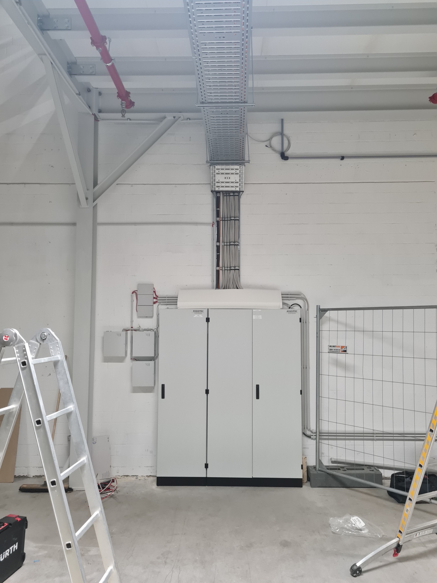 Rozdzielnia elektryczna zasilana wiązką kabli w korycie, widoczna w surowym, industrialnym wnętrzu, z rozłożonymi drabinami i narzędziami, zabezpieczona barierkami tymczasowymi.