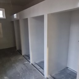 MIROS USŁUGI BUDOWLANE MYROSLAV MAKHOVSKYI - Wnętrze z wnękami wykończonymi na biało i szarą podłogą z płytek. Widoczne niedociągnięcia malarskie i wystające kable. Ujęcie z dołu, perspektywa podkreśla głębię pomieszczenia.