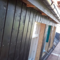 MIROS USŁUGI BUDOWLANE MYROSLAV MAKHOVSKYI - Czarna drewniana elewacja domu w trakcie montażu, widoczne okna zabezpieczone folią oraz fragment dachu z rynną.