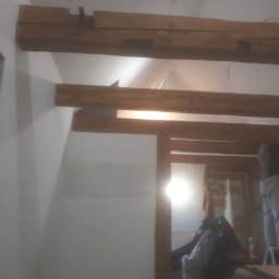 MIROS USŁUGI BUDOWLANE MYROSLAV MAKHOVSKYI - Wnętrze w trakcie remontu z widocznymi drewnianymi belkami stropowymi, częściowo pomalowanymi ścianami i zabezpieczonymi meblami folią ochronną.