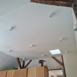 Biały sufit poddasza w Jeleniej Górze z drewnianymi belkami konstrukcyjnymi i punktowym oświetleniem LED. Widoczne okno dachowe i fragment pomieszczenia poniżej.