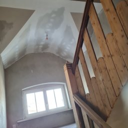 MIROS USŁUGI BUDOWLANE MYROSLAV MAKHOVSKYI - Poddasze w trakcie remontu: surowa ściana i skośny sufit z widocznymi łatami gipsu, drewniane schody z balustradą, okno w szpalecie z widokiem na zewnątrz.