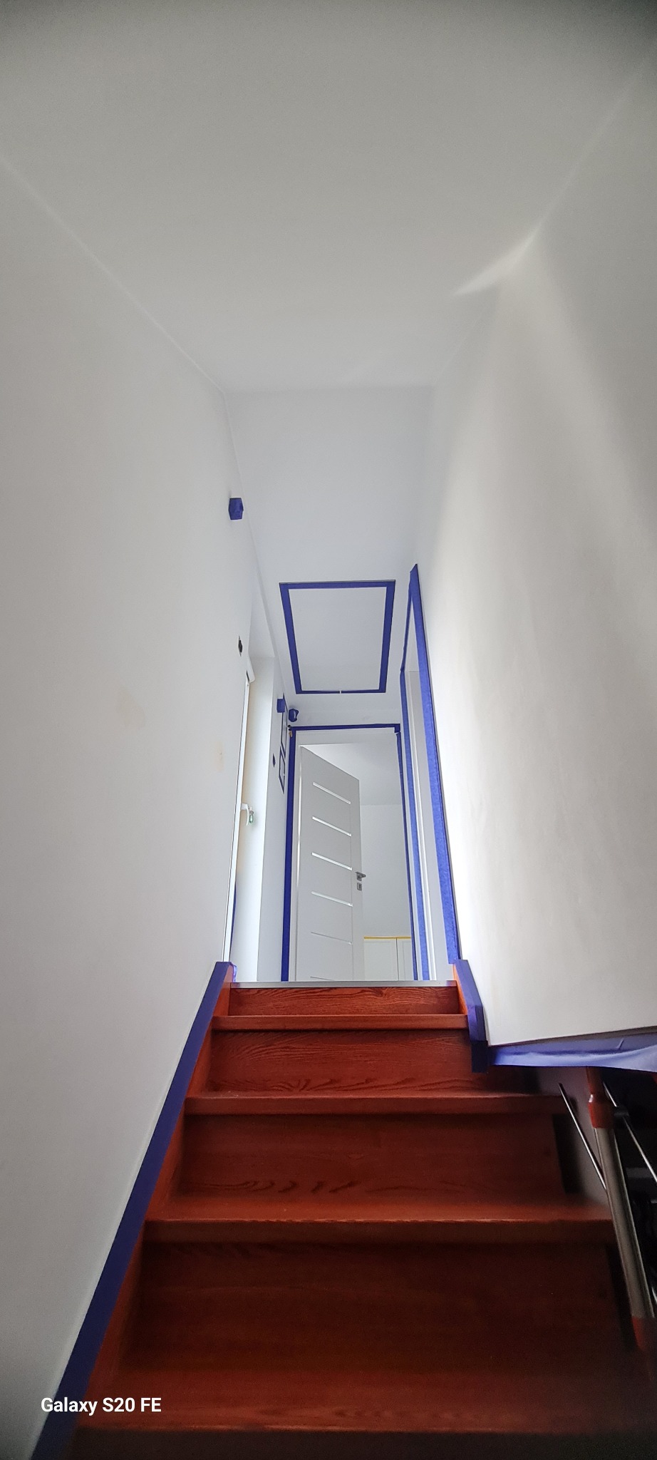 Klatka schodowa w trakcie malowania: białe ściany zabezpieczone niebieską taśmą malarską, drewniane schody. Widoczne otwarte drzwi i fragment sufitu.