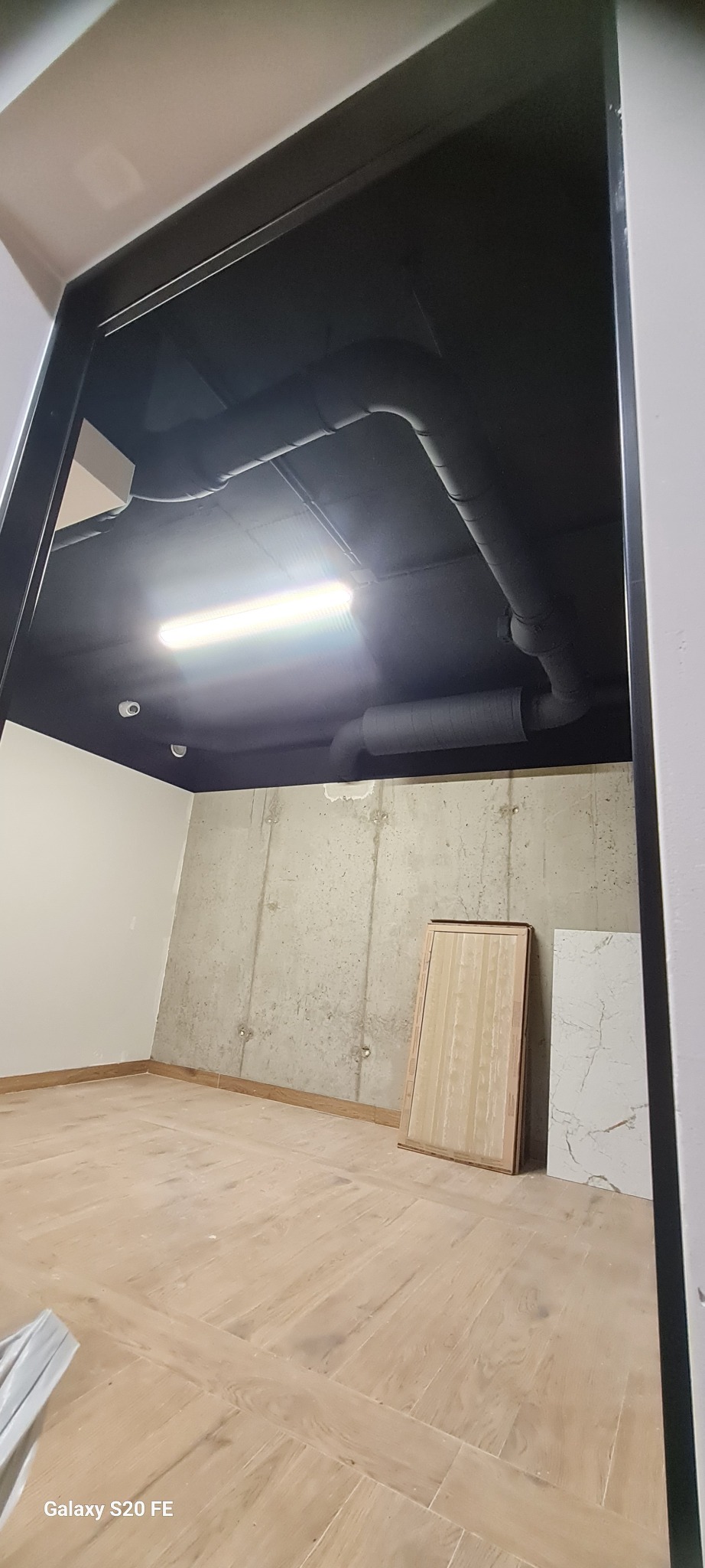 Surowy pokój z betonową ścianą, drewnianą podłogą i czarnym sufitem z rurami wentylacyjnymi. Widok zza framugi drzwi.