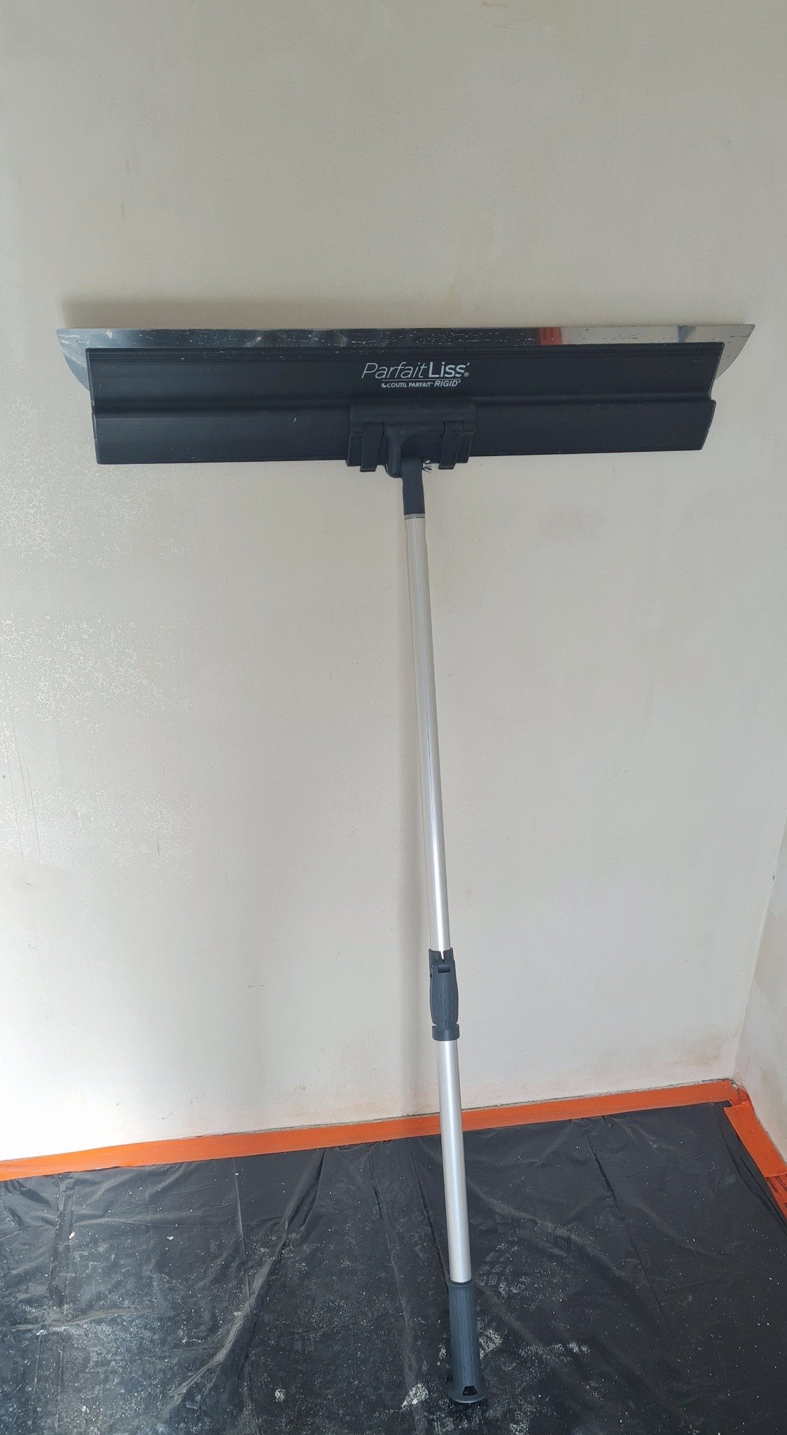 Narzędzie ParfaitLiss do gładzi na ścianie, z aluminiowym uchwytem. Podłoga zabezpieczona folią. Widoczny fragment ściany po nałożeniu gładzi.