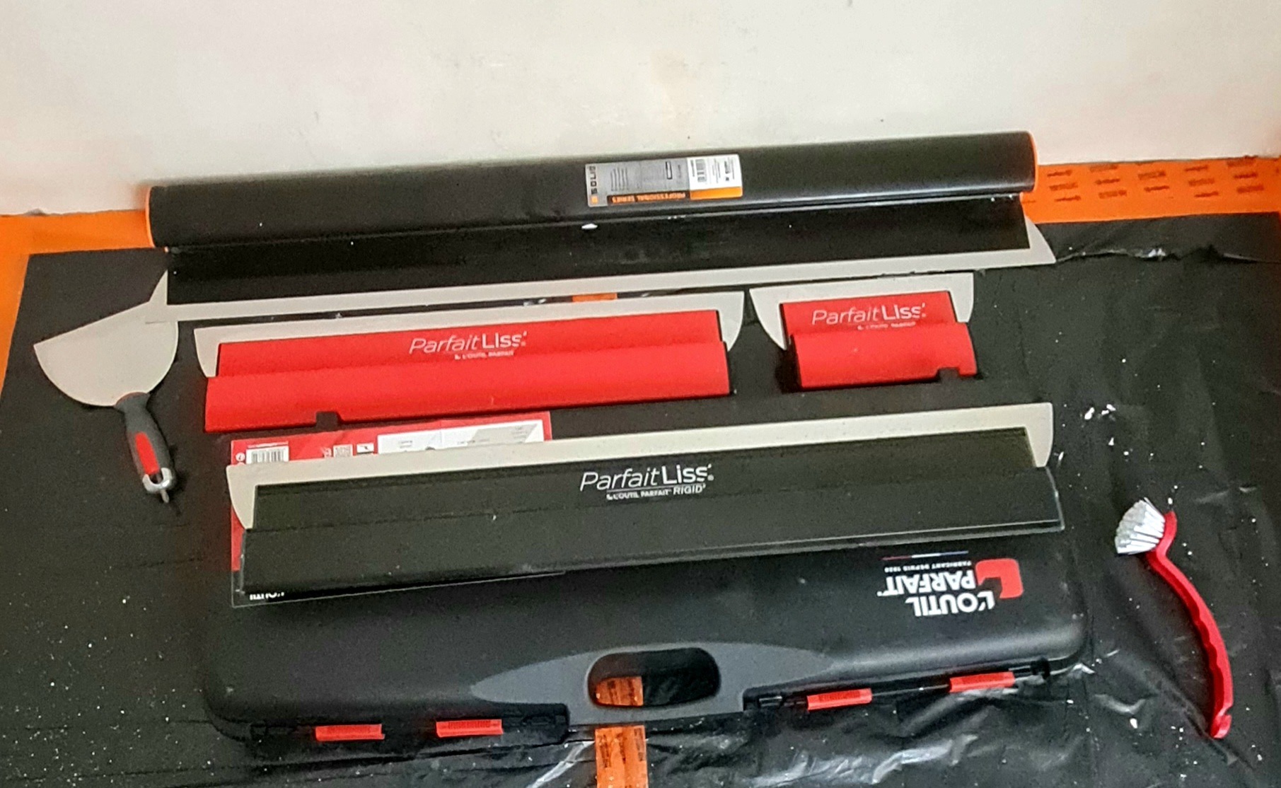 Zestaw narzędzi 'Parfait Liss' do gładzi szpachlowych: szpachelki, pędzle i kufer na czarnej folii ochronnej. Widoczne detale i logo producenta.