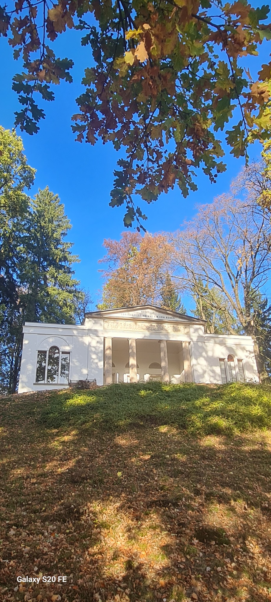 Klasycystyczna, biała budowla z kolumnami, widoczna z dołu, otoczona zielenią i drzewami z jesiennymi liśćmi na tle błękitnego nieba. Ujęcie z dołu podkreśla monumentalność.