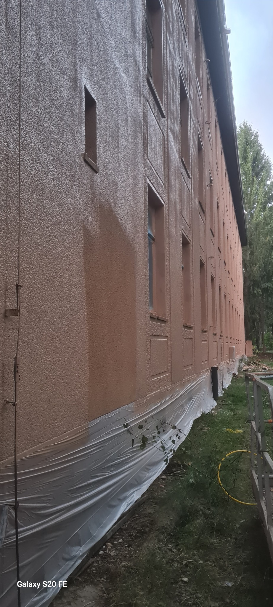 Świeżo pomalowana elewacja budynku wielorodzinnego w odcieniach brązu, zabezpieczona folią ochronną u podstawy. Widoczny fragment rusztowania i zieleń w tle.