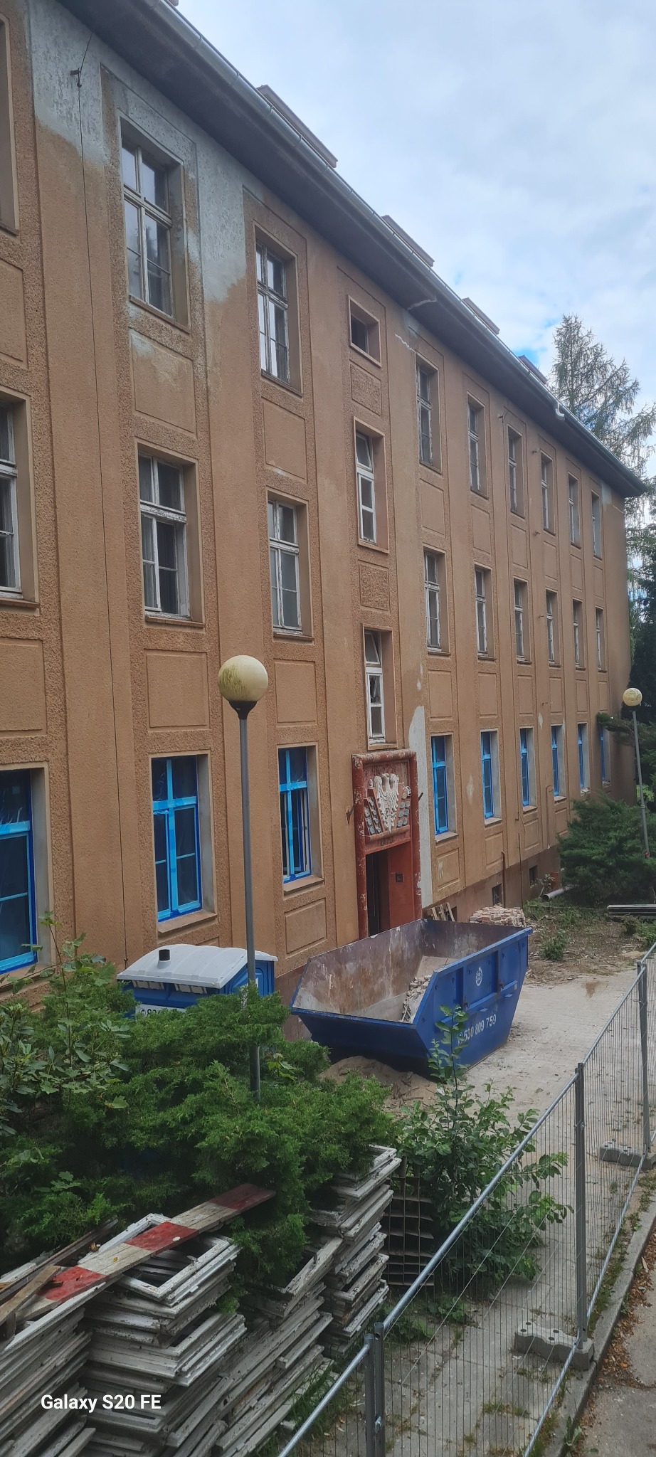 Budynek w trakcie prac elewacyjnych: częściowo ocieplony, okna zabezpieczone folią, kontener na gruz, stare okna składowane obok. Widok z dołu, lekki kąt.