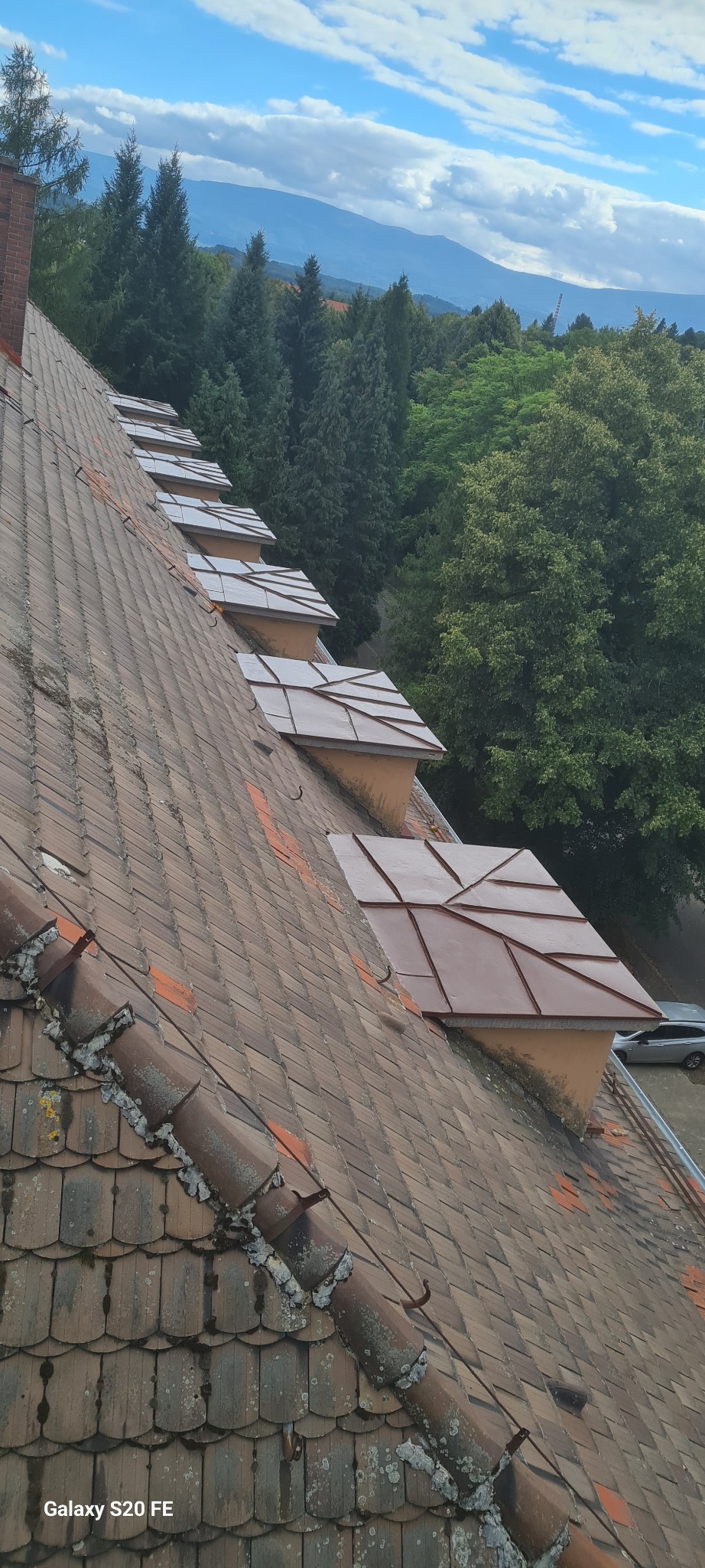 Dachówka karpiówka z nowymi lukarnami w Jeleniej Górze. Widok z góry na odnowiony dach z elementami blacharskimi. Tło stanowią drzewa i góry.