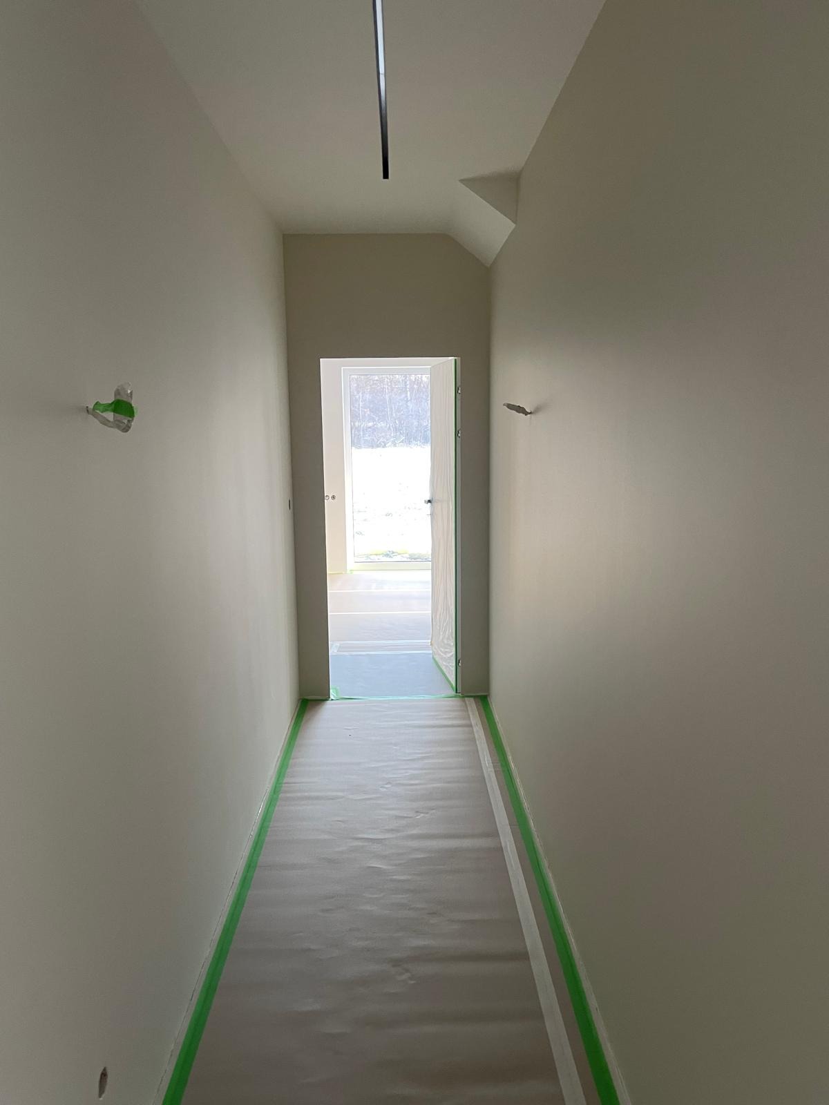 Korytarz w trakcie malowania: świeżo pomalowane ściany w jasnym odcieniu, zabezpieczona podłoga folią i taśmą malarską, widoczne ślady po szpachlowaniu.