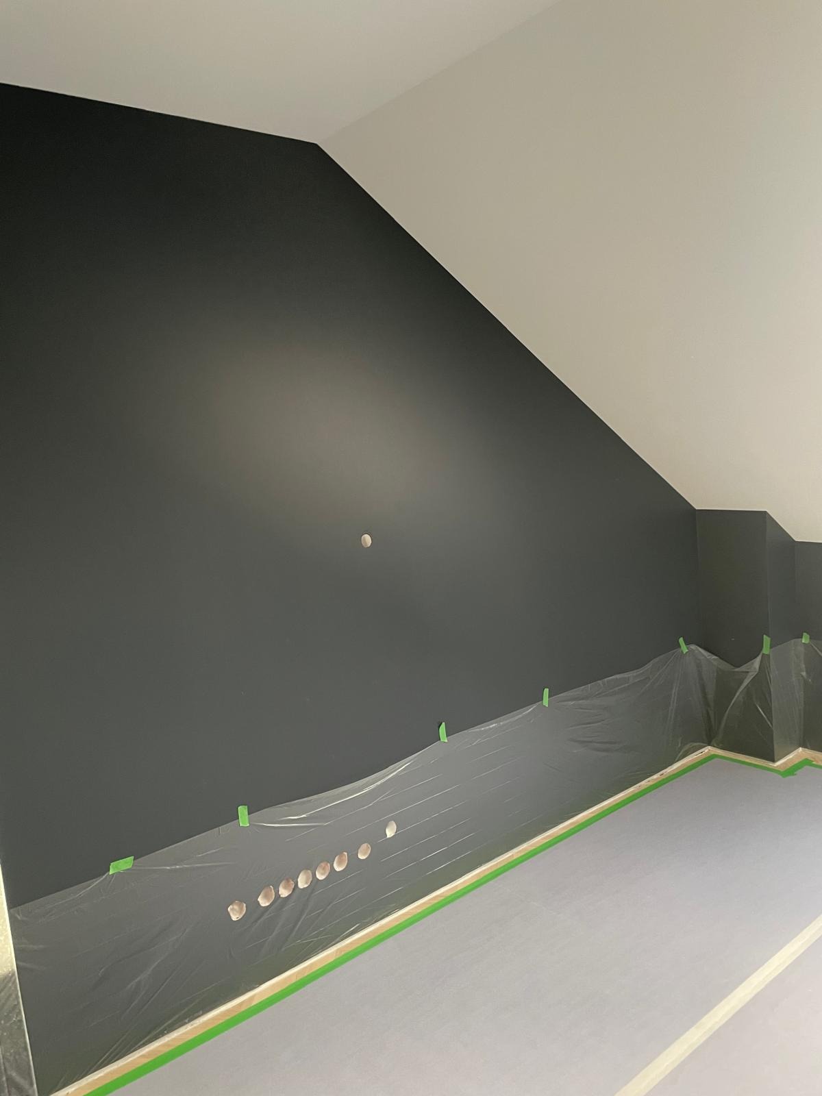 Ściana pomalowana na ciemny grafit w pokoju na poddaszu, zabezpieczona folią malarską i taśmą. Widoczne otwory na gniazdka elektryczne.