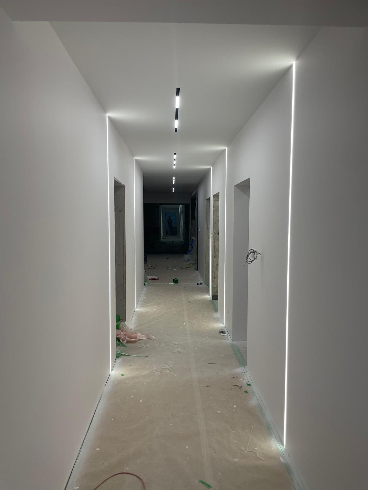 Długi korytarz po remoncie z minimalistycznym oświetleniem LED wzdłuż ścian i sufitu. Widoczne ślady prac wykończeniowych na podłodze.