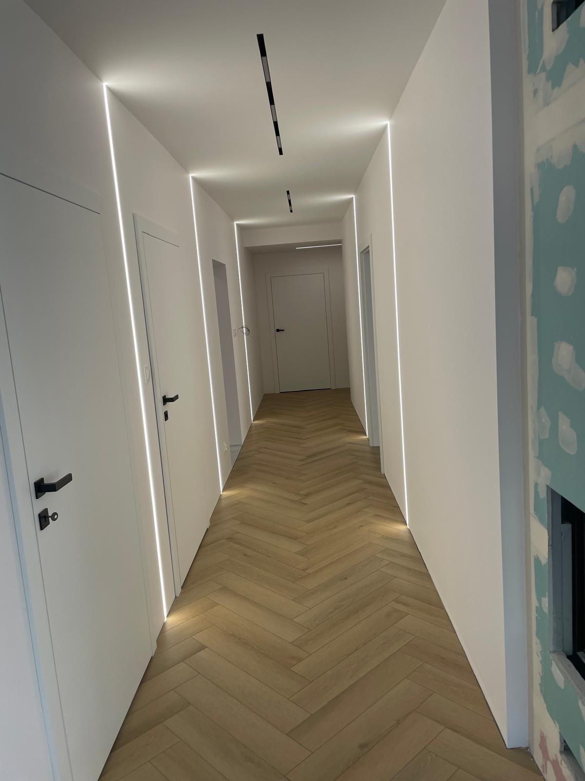 Nowoczesny korytarz po remoncie: białe ściany z oświetleniem LED, parkiet w jodełkę. Fragment ściany w trakcie wykończenia z płyt karton-gips.