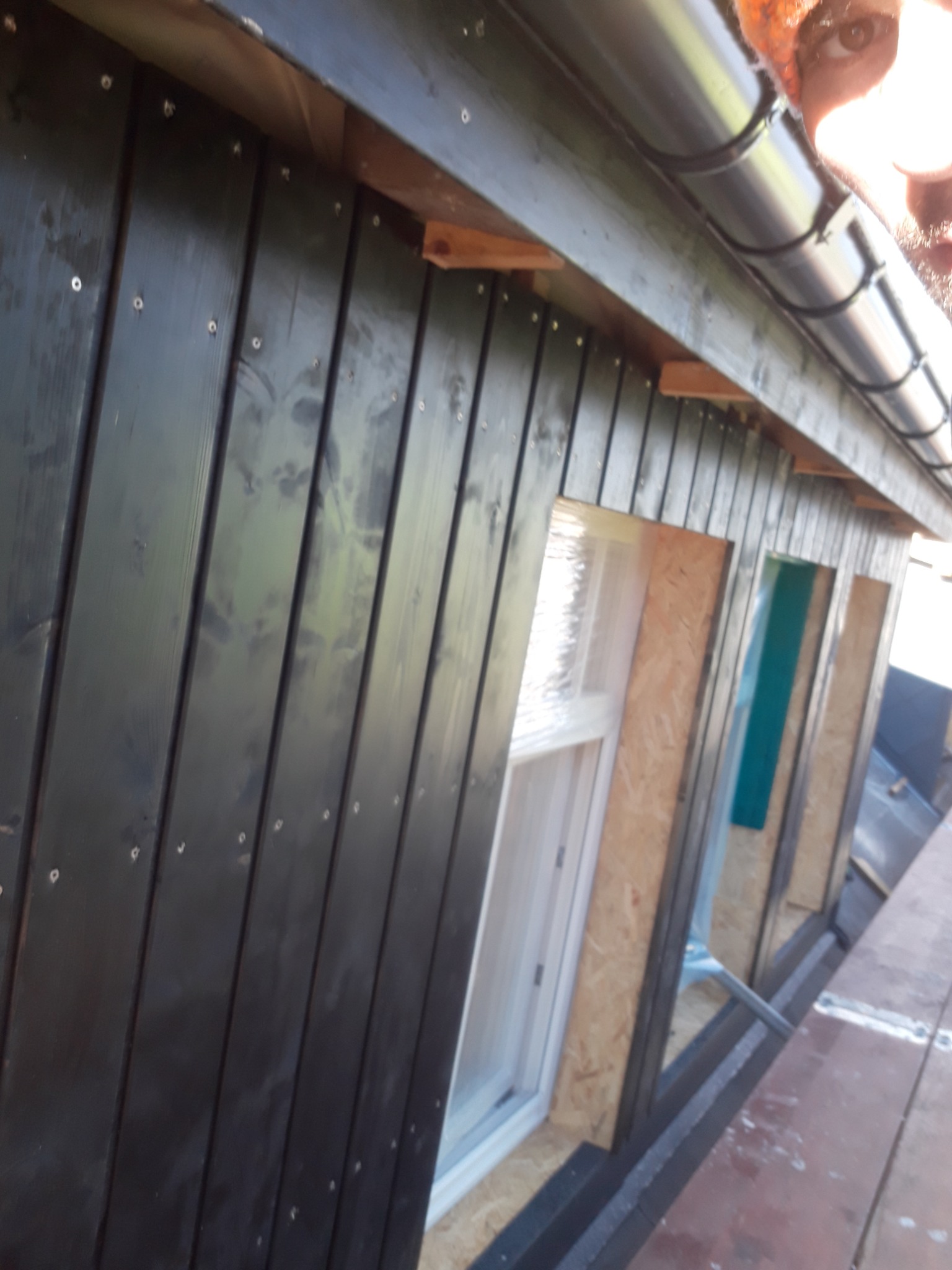 Czarna drewniana elewacja domu w trakcie montażu, widoczne okna zabezpieczone folią oraz fragment dachu z rynną.