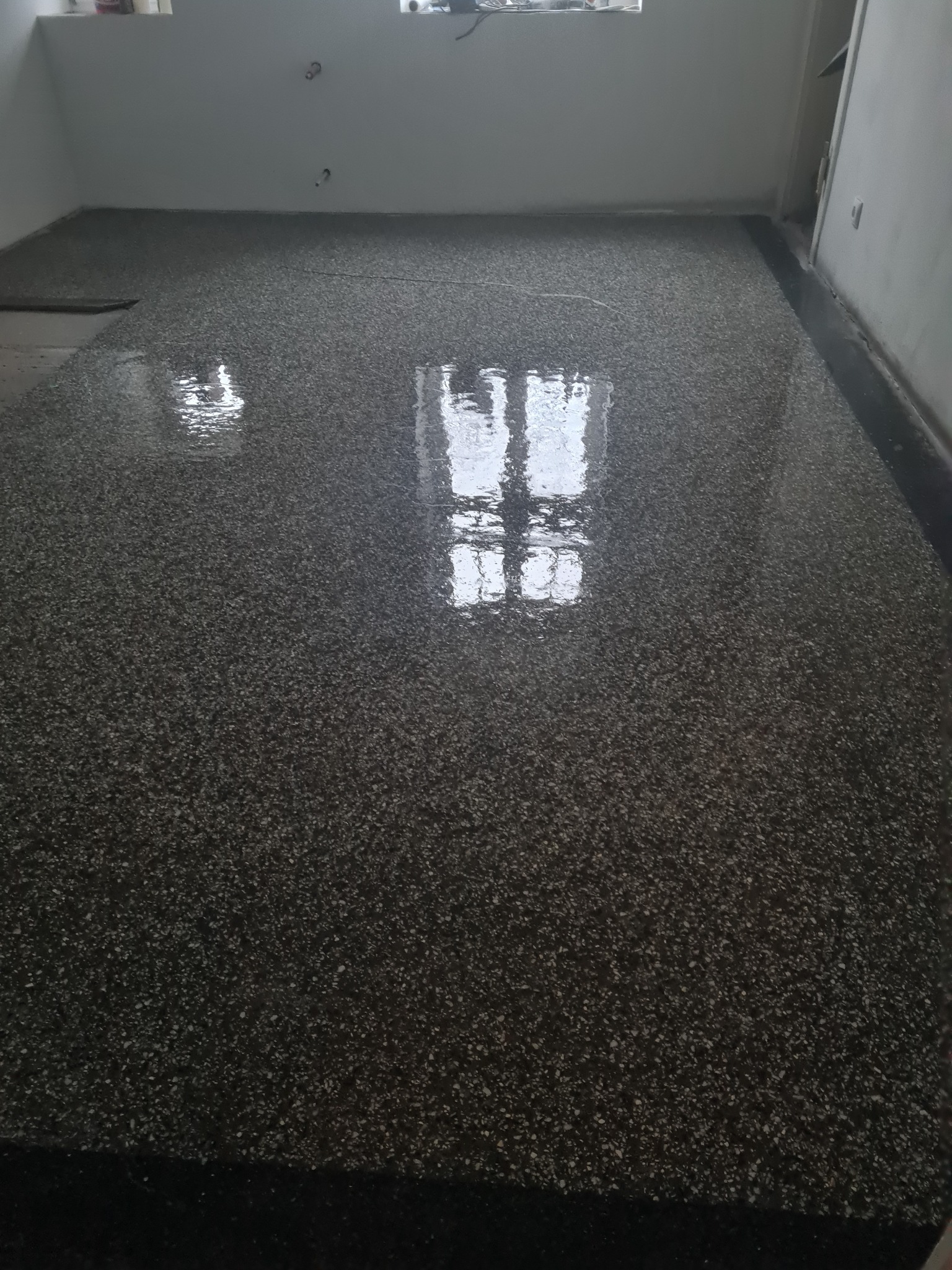 Świeżo wylana, błyszcząca, ciemna posadzka żywiczna z widocznym odbiciem okna, w surowym pomieszczeniu z białymi ścianami.