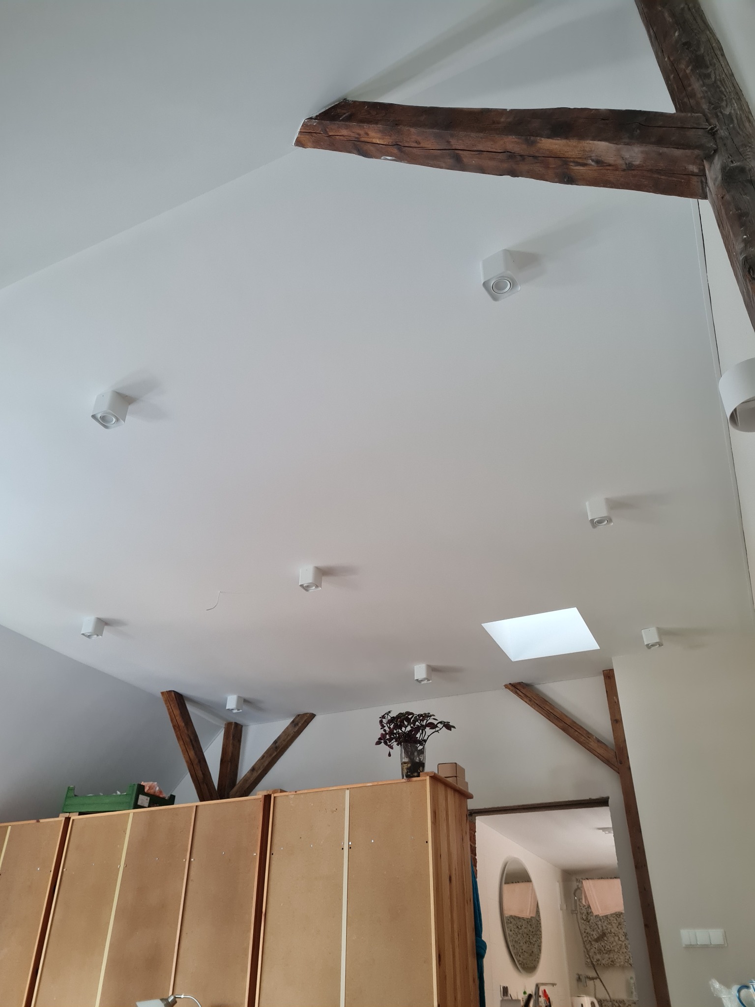 Biały sufit poddasza w Jeleniej Górze z drewnianymi belkami konstrukcyjnymi i punktowym oświetleniem LED. Widoczne okno dachowe i fragment pomieszczenia poniżej.