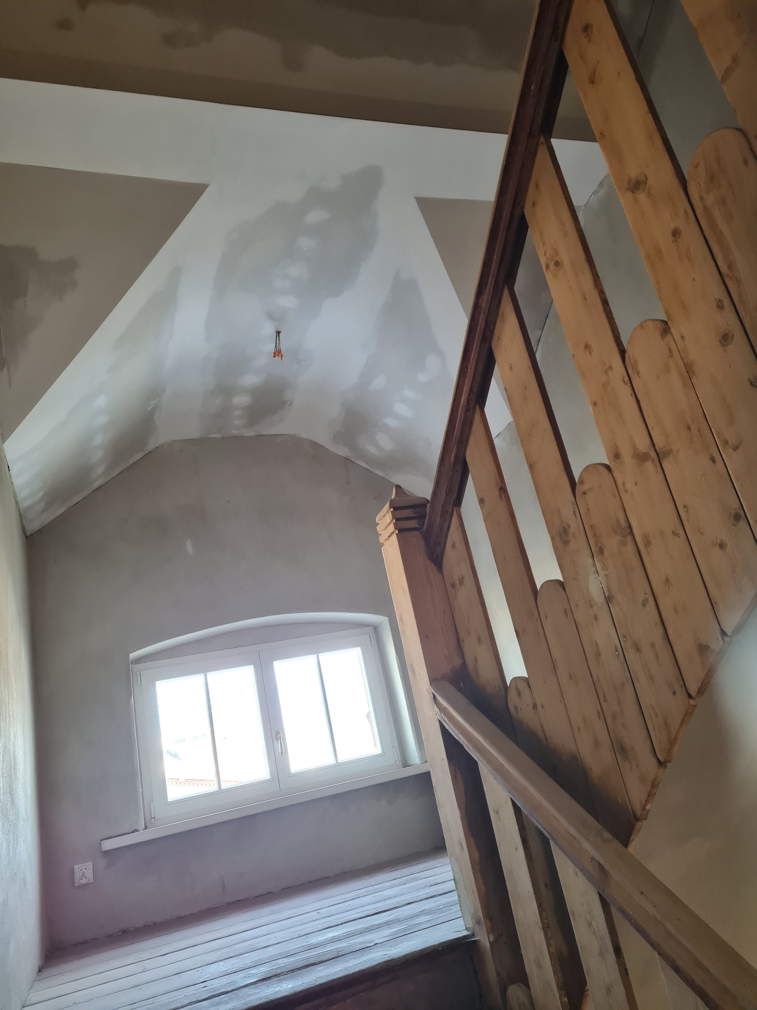 Poddasze w trakcie remontu: surowa ściana i skośny sufit z widocznymi łatami gipsu, drewniane schody z balustradą, okno w szpalecie z widokiem na zewnątrz.