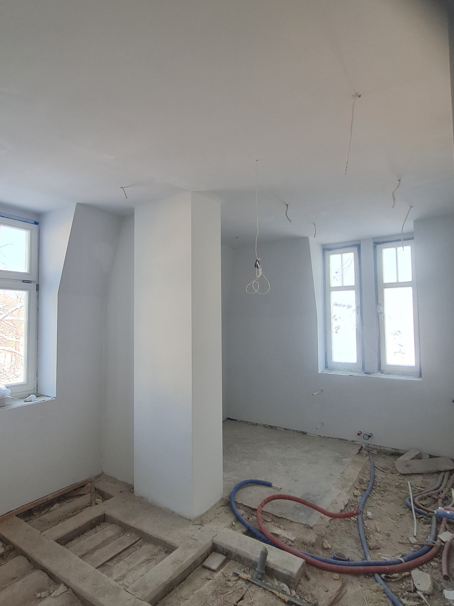 Wnętrze w trakcie remontu, świeżo pomalowane ściany na biało, widoczne instalacje elektryczne zwisające z sufitu, odkryta podłoga z widocznymi legarami i rurami instalacji wodnej.