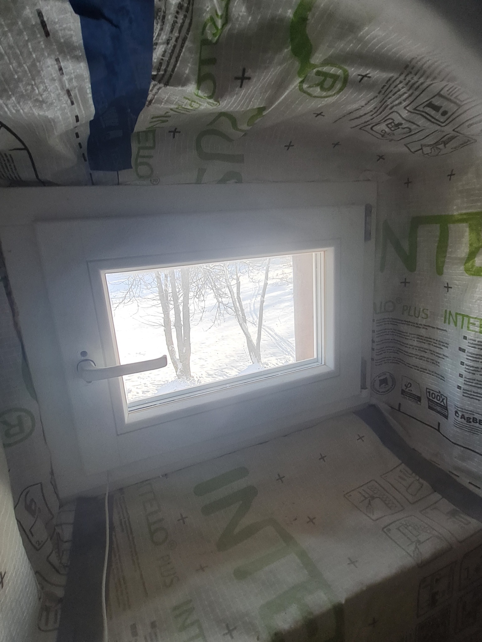 Wnętrze ocieplonego poddasza z widokiem na zimowy krajobraz przez białe okno PCV. Folia paroizolacyjna z nadrukiem widoczna na ścianach i suficie.