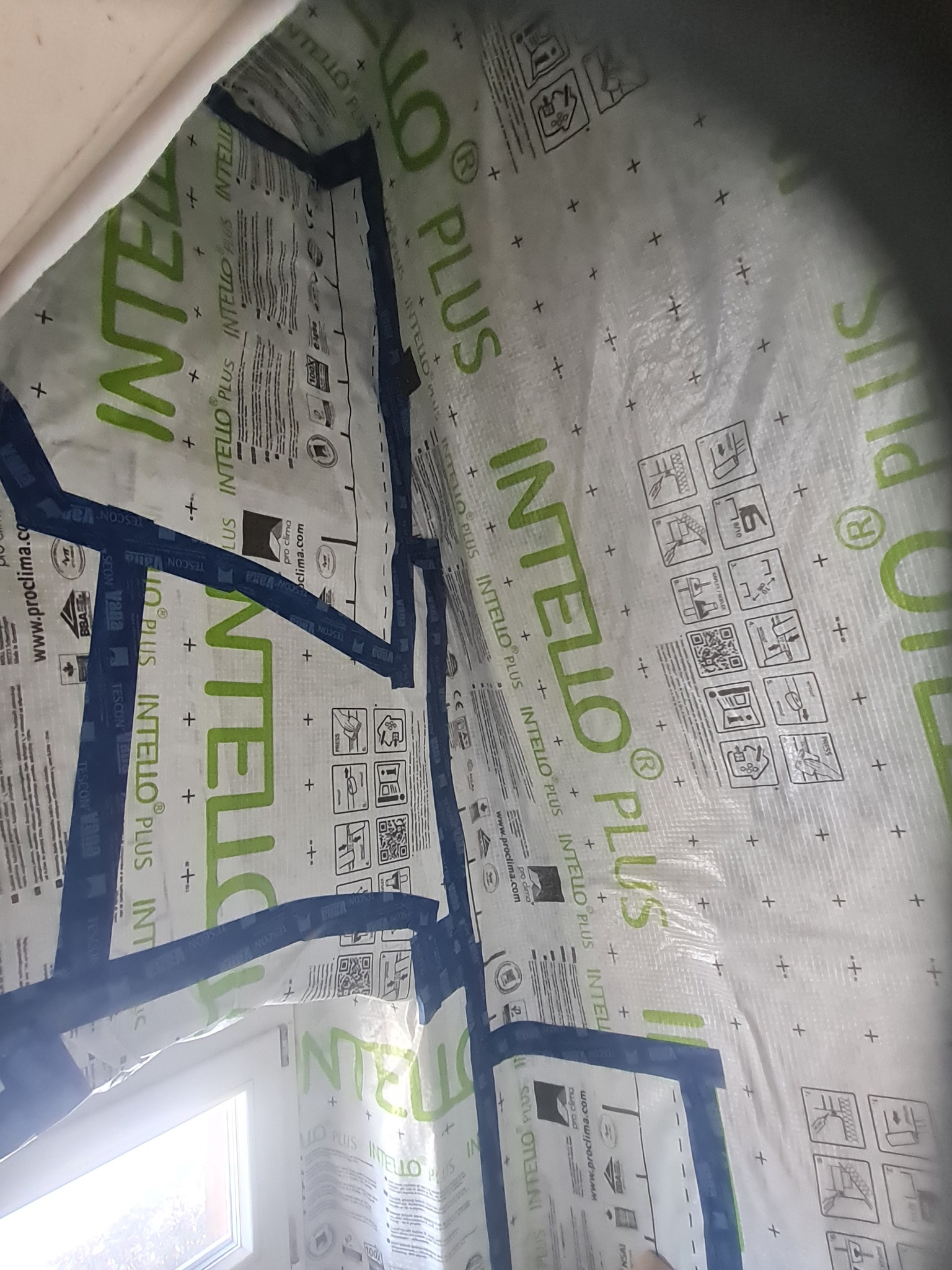 Szczegółowe ujęcie warstwy folii paroizolacyjnej Intello Plus, zabezpieczonej niebieską taśmą TESCON Vana, przytwierdzonej do krokwi poddasza, widok od wewnątrz, z fragmentem okna dachowego w tle.