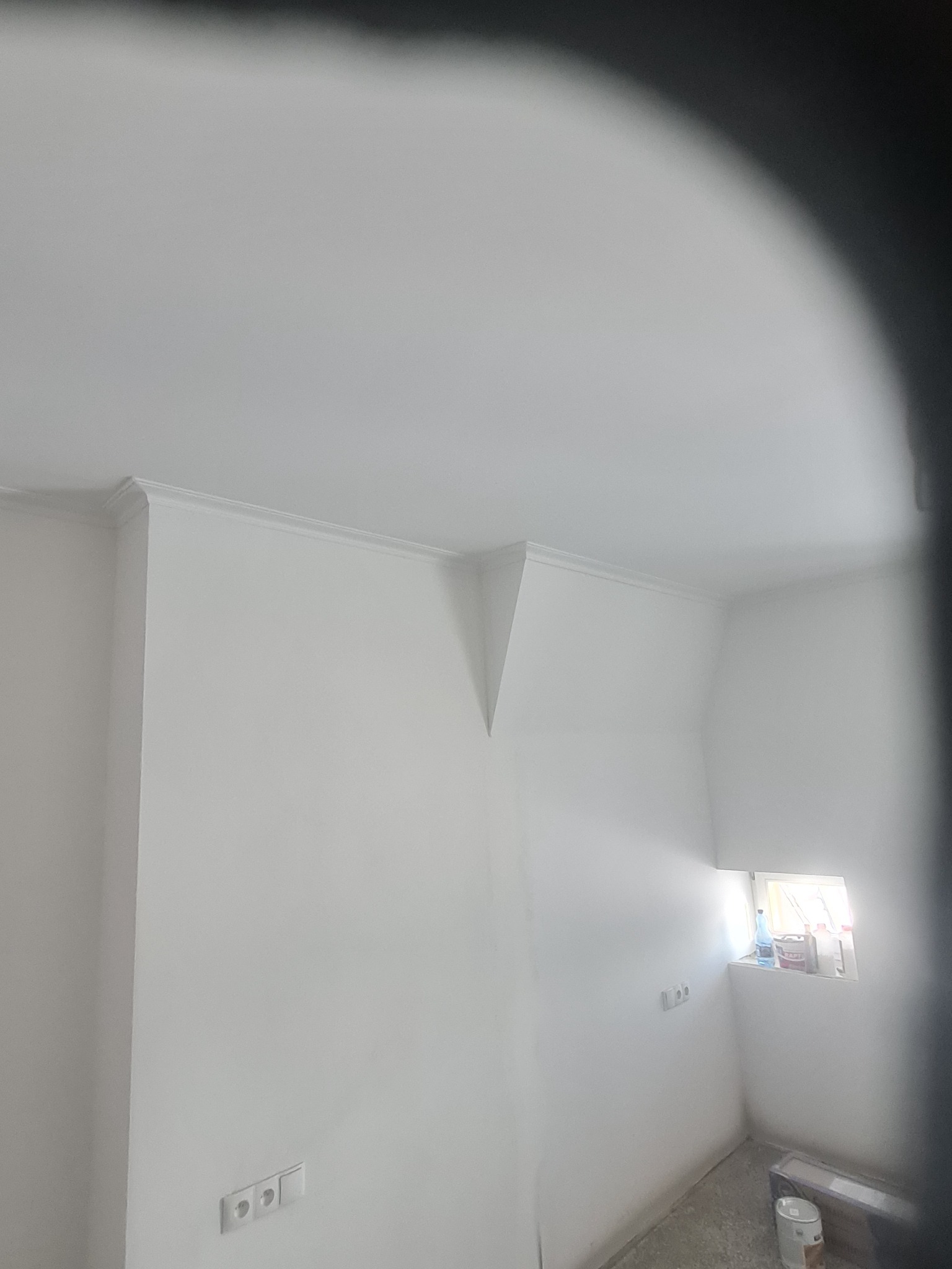 Wnętrze świeżo pomalowanego na biało pomieszczenia z listwami przysufitowymi, widoczne gniazdka elektryczne i okno z farbami na parapecie.