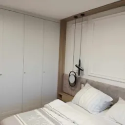 Jasna sypialnia z zabudowaną szafą wnękową o gładkich, białych frontach, nowoczesne oświetlenie wiszące i beżowe wezgłowie łóżka.