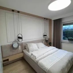 Nowoczesna sypialnia z szarą tapicerowaną ramą łóżka, podwieszanymi lampami z okrągłymi kloszami i dekoracyjną sztukaterią na ścianie.