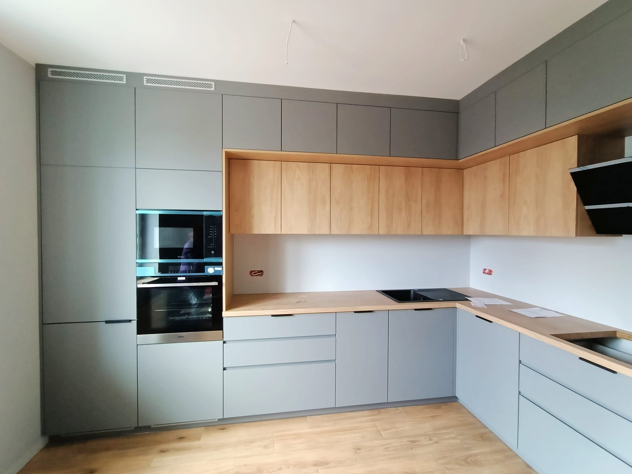 Nowoczesna kuchnia w odcieniach szarości i drewna z zabudowanym piekarnikiem i płytą grzewczą, widoczne elementy wentylacji pod sufitem i niezabezpieczone kable.