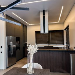 Stasacucine - Nowoczesna kuchnia STASACUCINE z ciemnymi, lakierowanymi szafkami, wyspą z drewnianym fornirem i oświetleniem LED w suficie, widziana z perspektywy stołu z wazonem suszonych kwiatów.