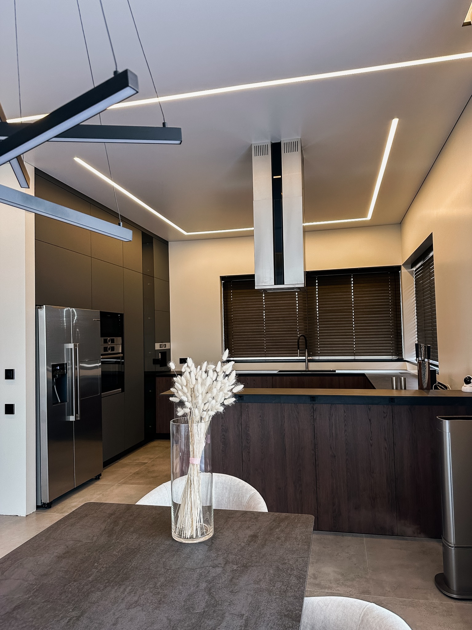Nowoczesna kuchnia STASACUCINE z ciemnymi, lakierowanymi szafkami, wyspą z drewnianym fornirem i oświetleniem LED w suficie, widziana z perspektywy stołu z wazonem suszonych kwiatów.