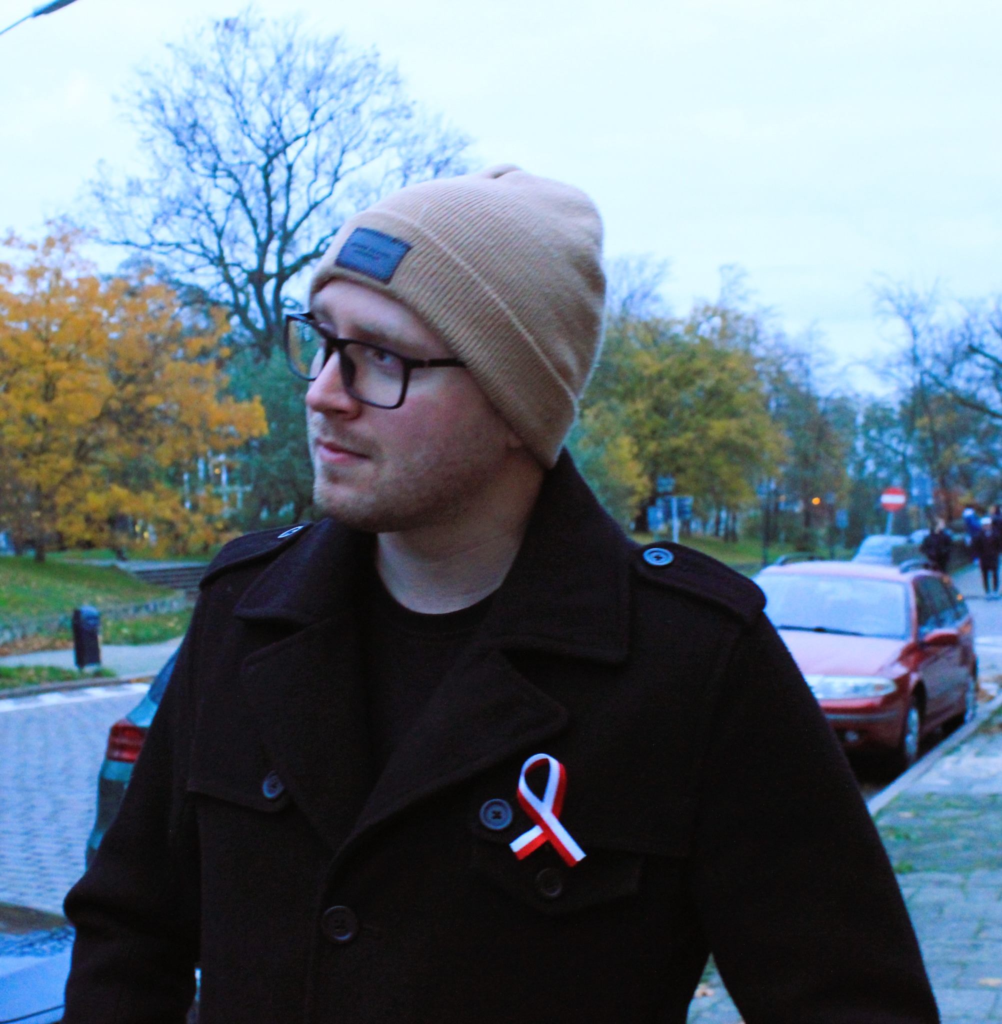 Mężczyzna w czarnym płaszczu z przypiętą biało-czerwoną kokardką, w beżowej czapce i okularach, patrzy w lewo na tle jesiennego krajobrazu miejskiego.