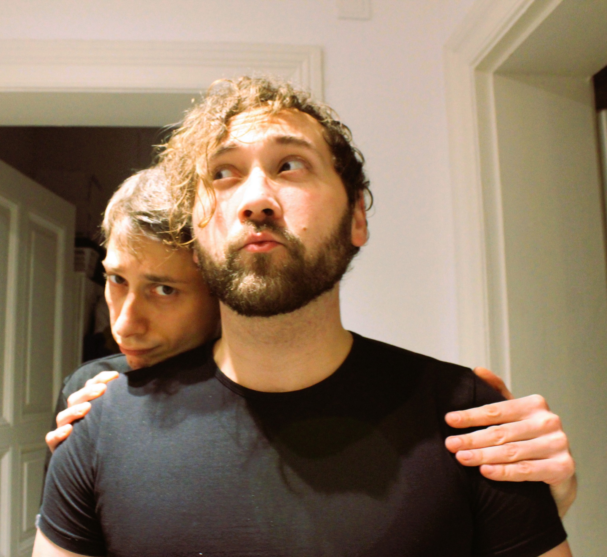 Portret dwóch mężczyzn w czarnych koszulkach na tle białej ściany i drzwi. Jeden z mężczyzn obejmuje drugiego ramieniem.