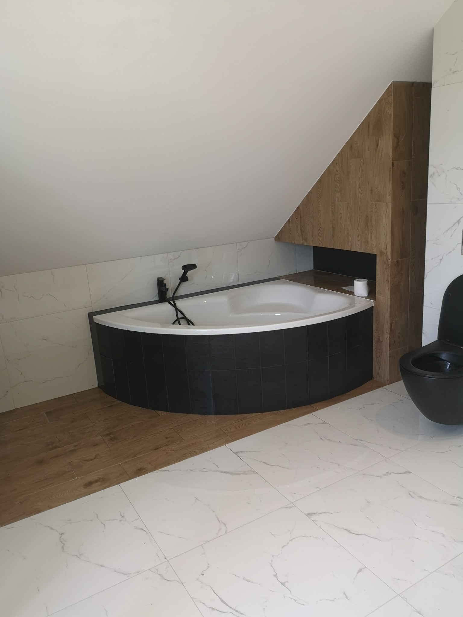 Nowoczesna łazienka na poddaszu z narożną wanną w czarnej obudowie, czarną armaturą i toaletą w tym samym kolorze, wykończona płytkami imitującymi marmur oraz drewnem.