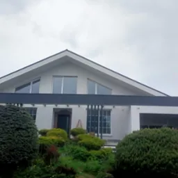 Nowoczesny dom jednorodzinny z białą elewacją, grafitowymi akcentami i dużymi oknami, otoczony bujną zielenią w pochmurny dzień.